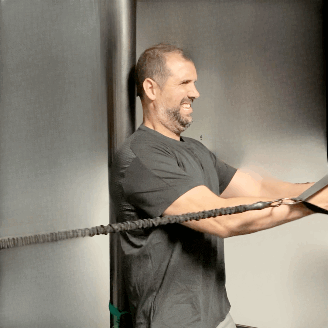 Man performing resistance band exercises while smiling, using a cable attachment anchored behind him for upper body training.