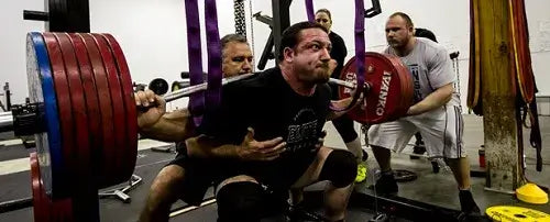 A group of lifters assisting a weightlifter squatting with a bent barbell loaded with weights.